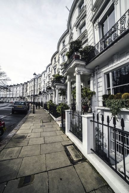 A street scene depicting a row of white, Victorian-style terraced houses with ornate iron balconies and decorative mouldings. The properties feature large windows, potted plants on the balconies and front steps, and black iron fencing along the pavement. A Bayswater Movers vehicle is partially visible, parked on the street, suggesting a home relocation process. The pavement is clean and wide, with few pedestrians and parked cars lining the road. The sky is overcast, casting diffused light over the scene, highlighting the architectural details and the orderly arrangement of the residential buildings. This setting reflects the typical surroundings encountered during furniture transport or packing and moving services within central London areas like Lancaster Gate, which are commonly covered in professional removals for house relocations. Bayswater Movers specializes in such services, supporting these detailed logistics in urban environments.