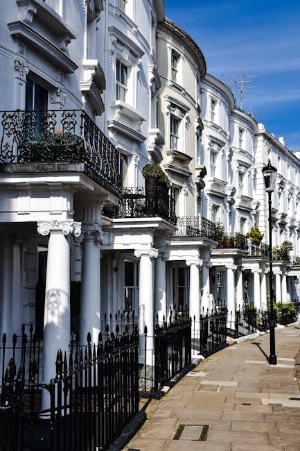 A row of white Victorian-style terraced houses with ornate architectural details, including decorative mouldings and curved bay windows, lining a clean, paved sidewalk under a bright blue sky. Each property features small, wrought-iron balconies with potted plants and black iron railings along the front. The street is partially shaded by a lamppost and a few trees, with a black metal fence separating the pavement from the front gardens. Inside one of the houses, a professional removal team from Bayswater Movers is engaged in a home relocation process, carefully loading furniture wrapped in plastic and blankets onto a moving van parked at the curb. Visible equipment includes a trolley and straps being used to facilitate the furniture transport, with cardboard boxes and packing materials nearby indicating a packing and moving operation. The scene exemplifies typical house removal activities, highlighting the logistical steps involved in furniture transport during relocation services. This image is relevant to the Lancaster Gate removals insider tips for Bayswater, showcasing the detailed process of house removals in a residential street.