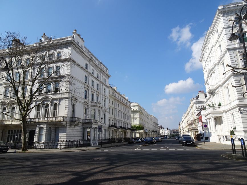 A street scene depicting a row of white, Victorian-style terraced houses with ornate iron balconies and decorative mouldings. The properties feature large windows, potted plants on the balconies and front steps, and black iron fencing along the pavement. A Bayswater Movers vehicle is partially visible, parked on the street, suggesting a home relocation process. The pavement is clean and wide, with few pedestrians and parked cars lining the road. The sky is overcast, casting diffused light over the scene, highlighting the architectural details and the orderly arrangement of the residential buildings. This setting reflects the typical surroundings encountered during furniture transport or packing and moving services within central London areas like Lancaster Gate, which are commonly covered in professional removals for house relocations. Bayswater Movers specializes in such services, supporting these detailed logistics in urban environments.