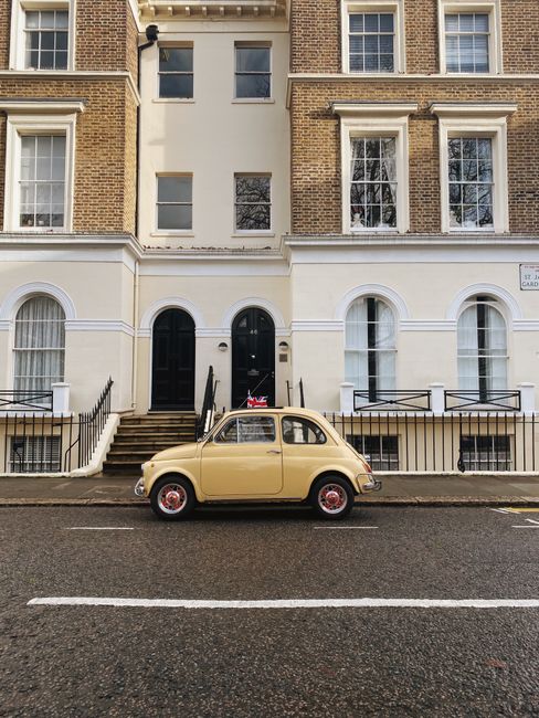 A narrow street with a row of terraced houses featuring brick and white facades, large windows, and decorative window framing. In front, a beige vintage car is parked parallel to the pavement, which is wet, indicating recent rain. The house at the center has a black door with a small window and a Union Jack flag attached near the entrance. On the steps leading up to the doors are black metal railings. A cardboard box, plastic wrapping, and a moving blanket are visible on the pavement in front of the house, suggesting a home relocation or furniture transport process. This scene aligns with professional removals services, such as those provided by Bayswater Movers, especially in the context of navigating narrow Bayswater streets for house removals and packing and moving activities.