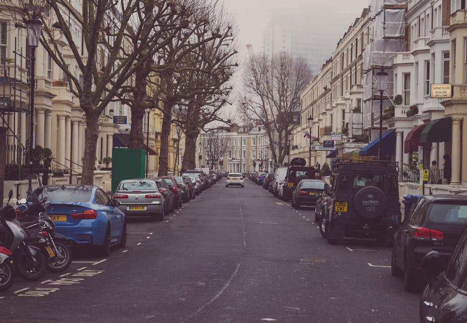 A narrow residential street in Bayswater during daytime with parked cars lining both sides, including a blue sports car, a silver sedan, and a black off-road vehicle with a spare tire on the back. Several motorcycles are parked on the pavement near the blue car. The street is bordered by white Victorian-style terraced houses with large windows, some featuring decorative columns and ornate balconies. Tall leafless trees are planted along the curb, casting shadows on the asphalt. In the background, a white van is seen reversing or stationary further down the street. The scene appears overcast, with diffused natural light showcasing the urban environment typical of home relocations and furniture transport logistics, as indicated by the [COMPANY_NAME] on some vehicles and the context of house removals in the BAYSWATER area.